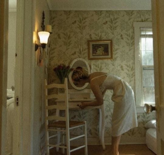 A woman in a white slip leaning over a small white dressing table in a room papered with leaves, a lamp lit on the wall.