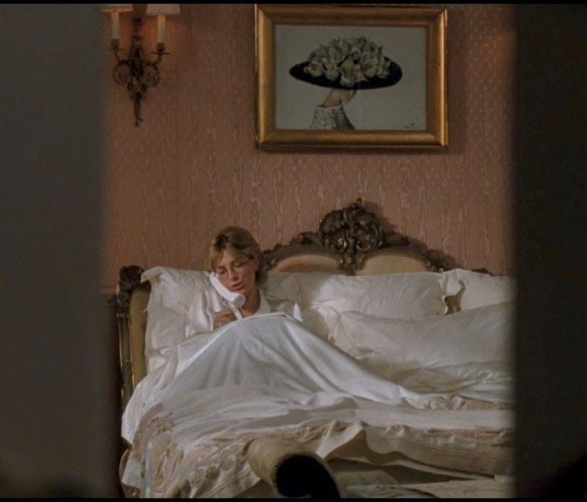 A still from The Parent Trap: a woman sitting up in a white bed in a pink bedroom, writing, under a painting of a hat.